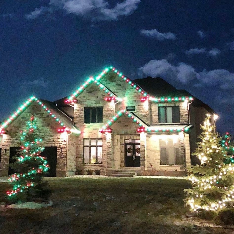 Large stone home with red and green roofline lights and red and green wrapped trees